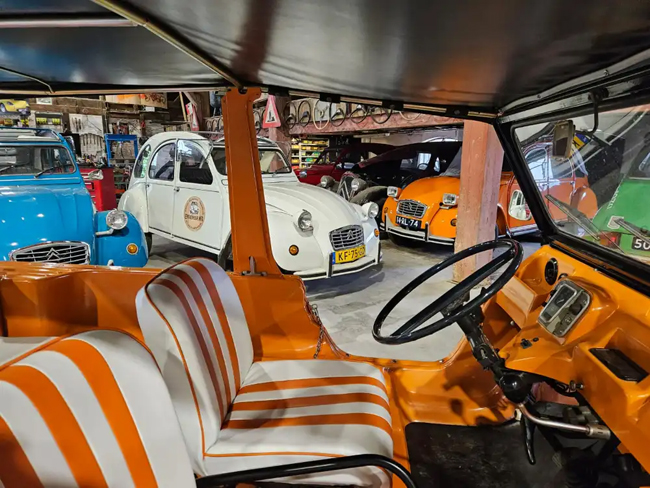 Interieur van een oranje Citroën Mehari in een garage met een verzameling Citroën 2CV auto's op de achtergrond